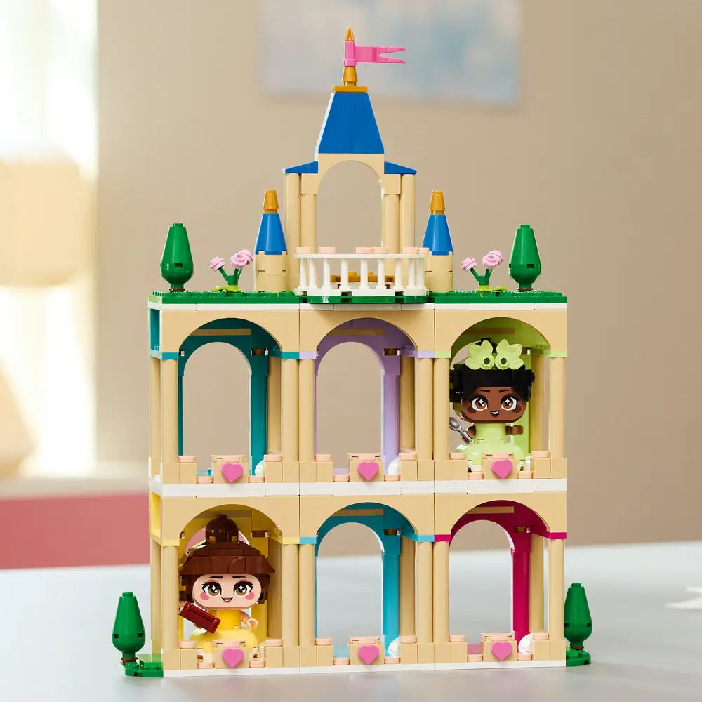 LEGO Disney Princess Mini Belle and Tiana Castle fully built, featuring tan columns, pastel archways, blue spires, balcony railings, and character figures displayed against a softly lit room backdrop.