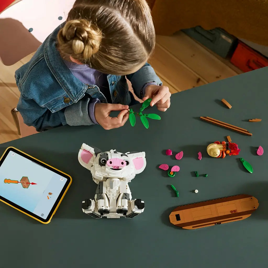 LEGO Disney Moana Pua set in white, gray, pink, green, and brown plastic is assembled by a child at a dark table beside tablet instructions, loose pieces, Heihei, and a brown base.