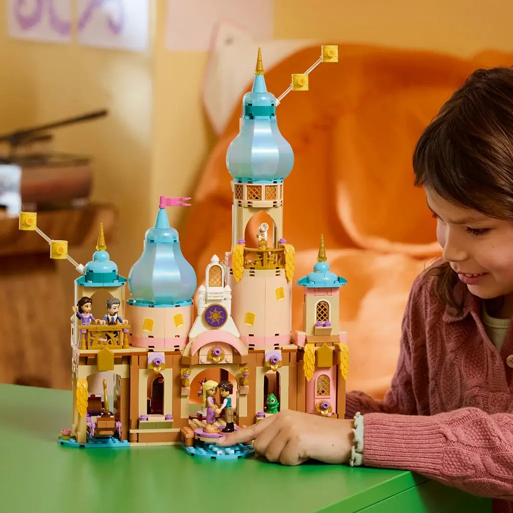 LEGO Disney Princess Rapunzel's Castle in tan, pink, turquoise, and gold plastic stands on a green table as a child plays with figures at the palace entrance and turntable.