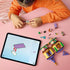 LEGO Disney Princess Rapunzel's Mini Tower pieces in yellow, tan, purple, and green plastic cover a pink table as a child builds the hinged tower beside tablet instructions.
