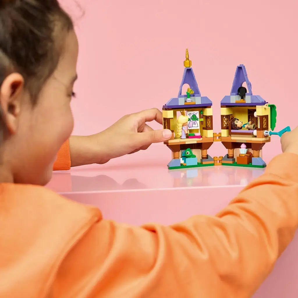 LEGO Disney Princess Rapunzel's Mini Tower in yellow, tan, purple, and green plastic is opened to reveal rooms as a child plays with Rapunzel and Flynn Rider on a pink table.