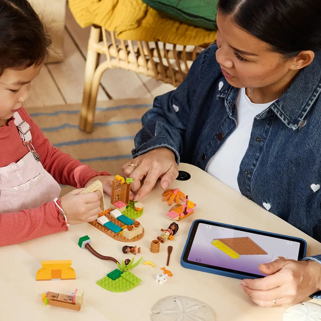LEGO Disney Moana Village House & Boat in colorful plastic pieces is built at a table by an adult and child using tablet instructions, with figures and parts spread nearby.