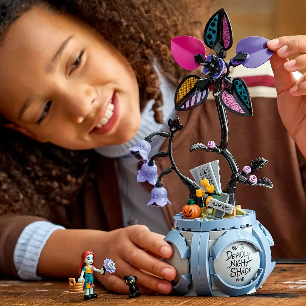 LEGO Disney Sally flowerpot toy features a blue plastic cauldron base with black twisting stems and bright pink, purple, teal, and yellow petals, held by a child while assembling the spooky themed flower in a cozy indoor setting.
