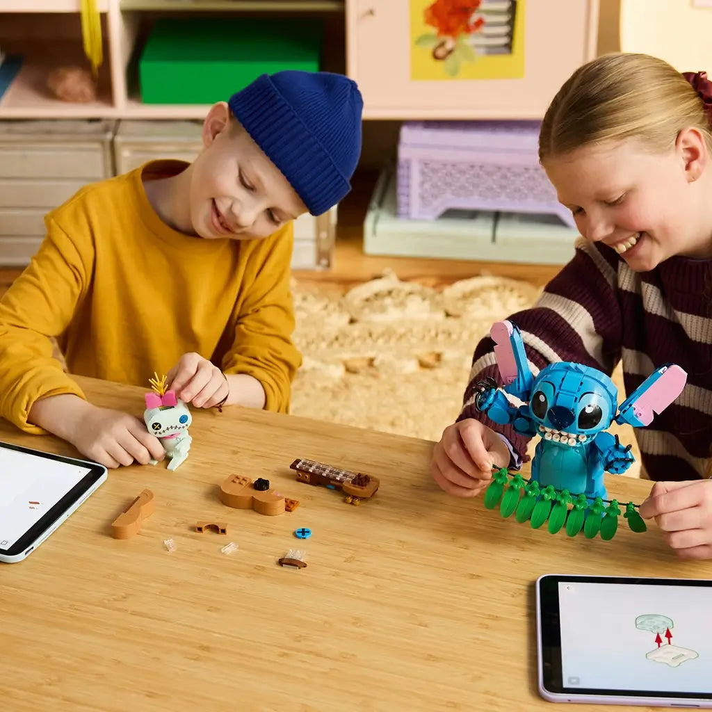 LEGO Disney Stitch and Scrump set in blue, pink, green, and tan plastic is built by two smiling children at a wooden table with loose guitar pieces and tablets.