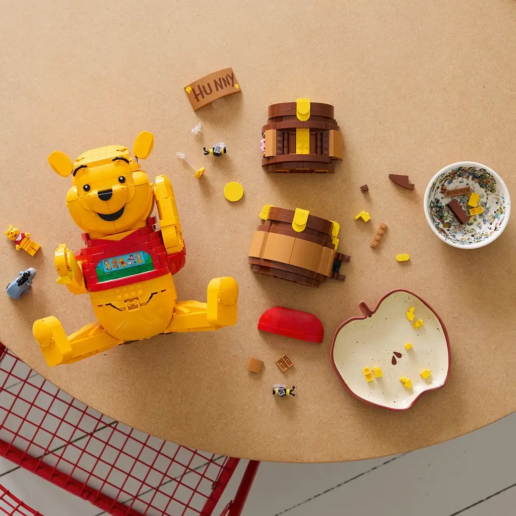 LEGO Disney Winnie the Pooh building set in yellow, red, and brown plastic pieces, disassembled figure and honey pot components, creative construction scene on a tabletop workspace.