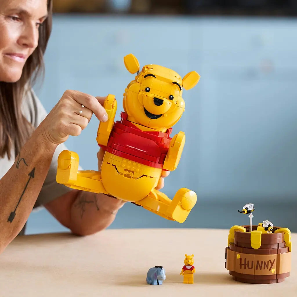 LEGO Disney Winnie the Pooh build in yellow and red plastic, rounded bear model being assembled by adult hands, interactive display toy with honey pot and figures on a tabletop setting.