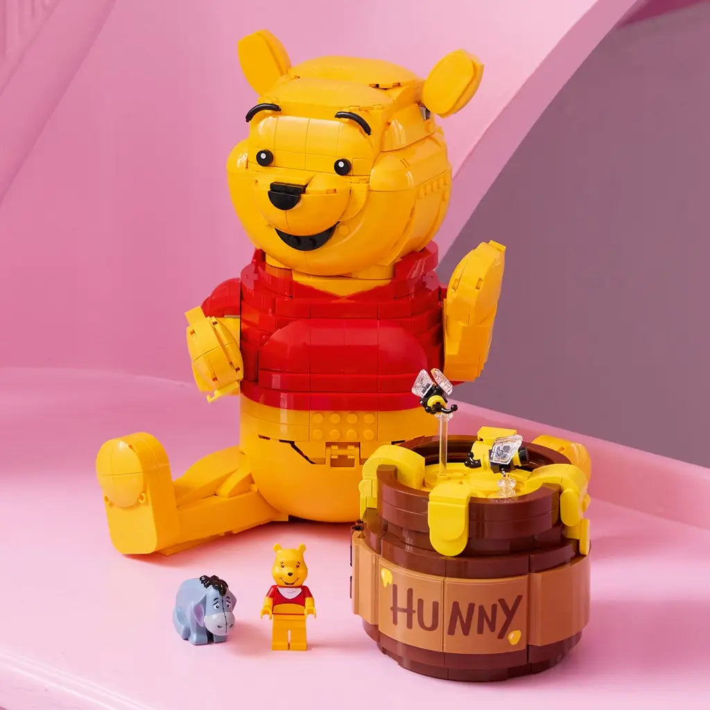 LEGO Disney Winnie the Pooh display model in yellow and red plastic, seated bear figure with honey pot and mini characters, decorative toy arranged on a pastel pink background.