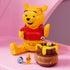 LEGO Disney Winnie the Pooh display model in yellow and red plastic, seated bear figure with honey pot and mini characters, decorative toy arranged on a pastel pink background.