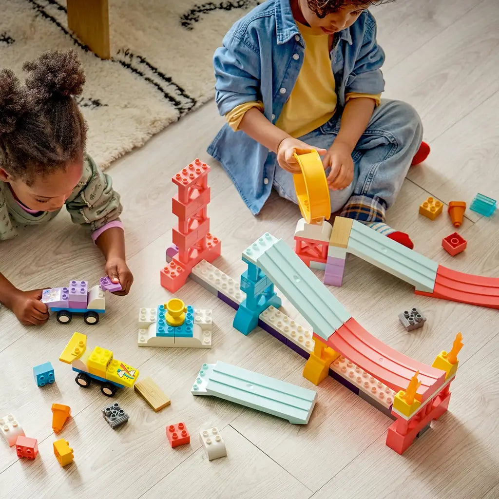 LEGO DUPLO Creative Ramps playset features pastel plastic ramps, towers and an orange loop as two children build and race colorful cars together on a wooden floor.