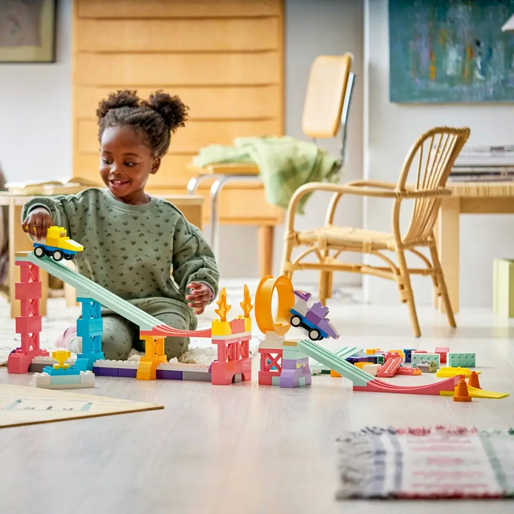 LEGO DUPLO ramp playset features pastel plastic towers, slopes and an orange loop as a child races yellow and purple cars across a bright living room floor.