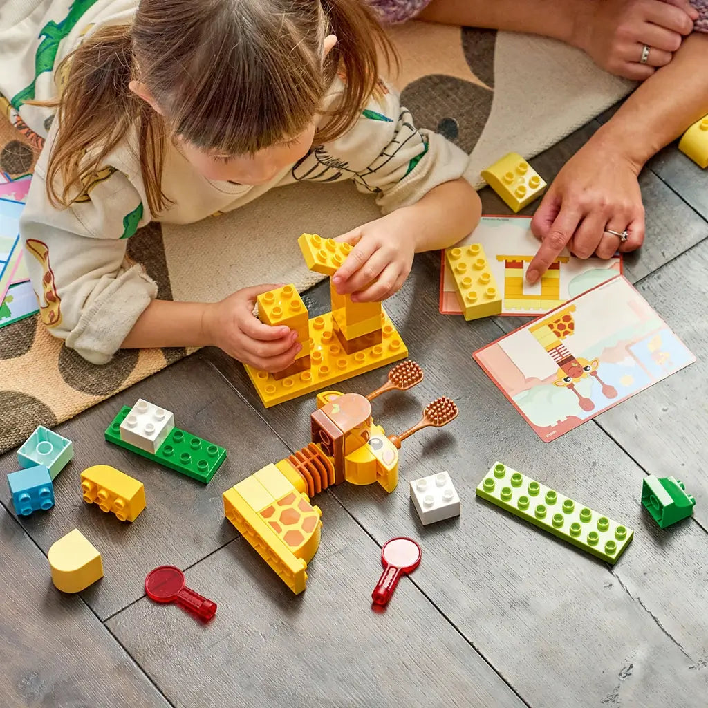 LEGO DUPLO Animal Building Game play scene shows a child stacking yellow plastic bricks for a giraffe while an adult points to a matching picture card on a wood floor.
