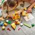 LEGO DUPLO Animal Building Game play scene shows a child stacking yellow plastic bricks for a giraffe while an adult points to a matching picture card on a wood floor.