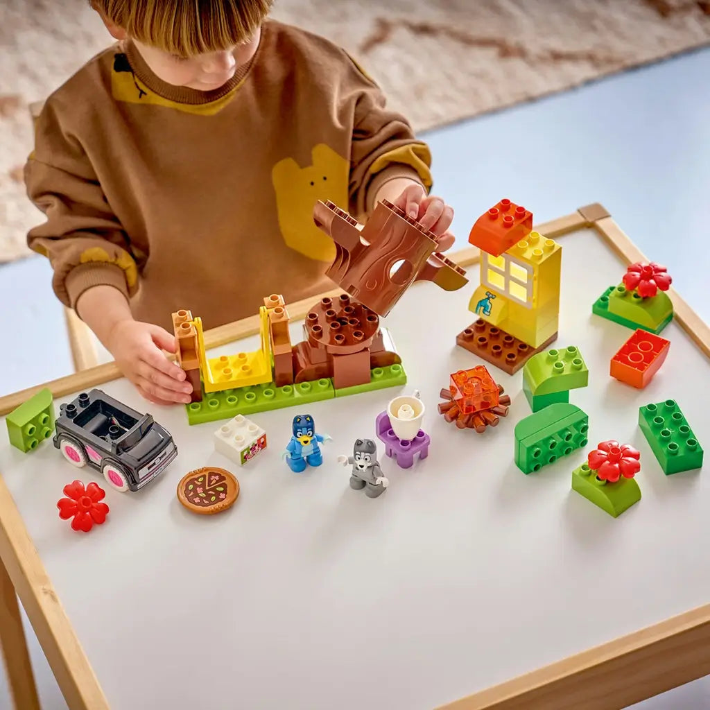 LEGO DUPLO Bluey Pizza Girls playset uses colorful plastic bricks as a child builds a brown tree beside a house, campfire, car, pizza, Bluey and Muffin figures.