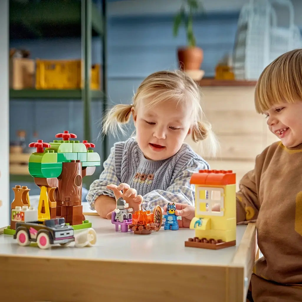 LEGO DUPLO Bluey Pizza Girls playset shows colorful plastic tree, house, campfire, car and character figures as two children play together at a wooden table indoors.