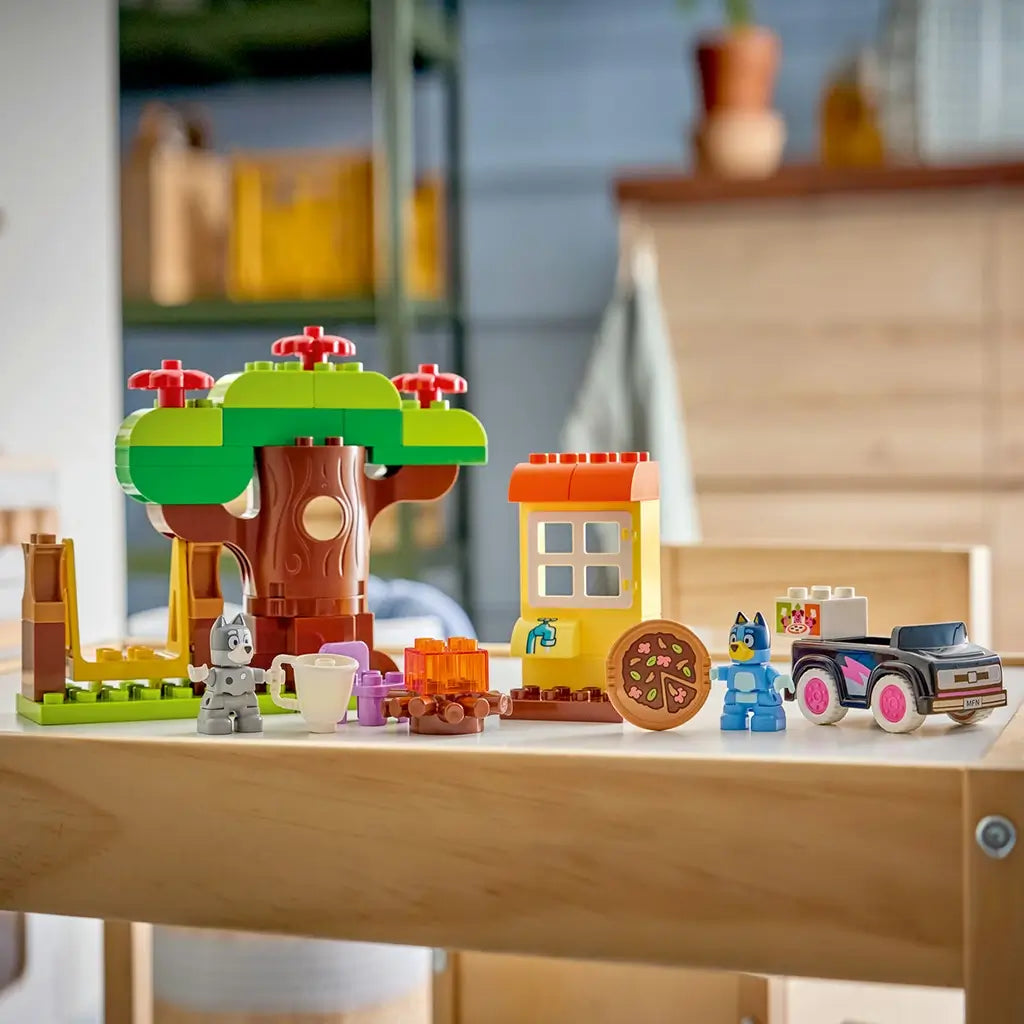 LEGO DUPLO Bluey Pizza Girls playset features colorful plastic tree, house, campfire, pizza, car, Bluey and Muffin figures arranged on a wooden table indoors.