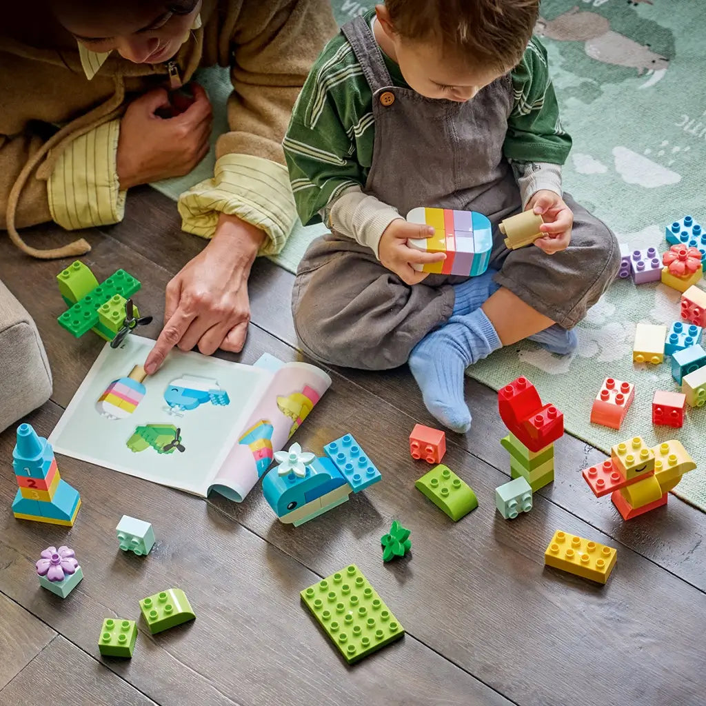 LEGO DUPLO Colorful Creative Box bricks in bright plastic colors surround a child building a striped model while an adult points to ideas in a book on the floor.