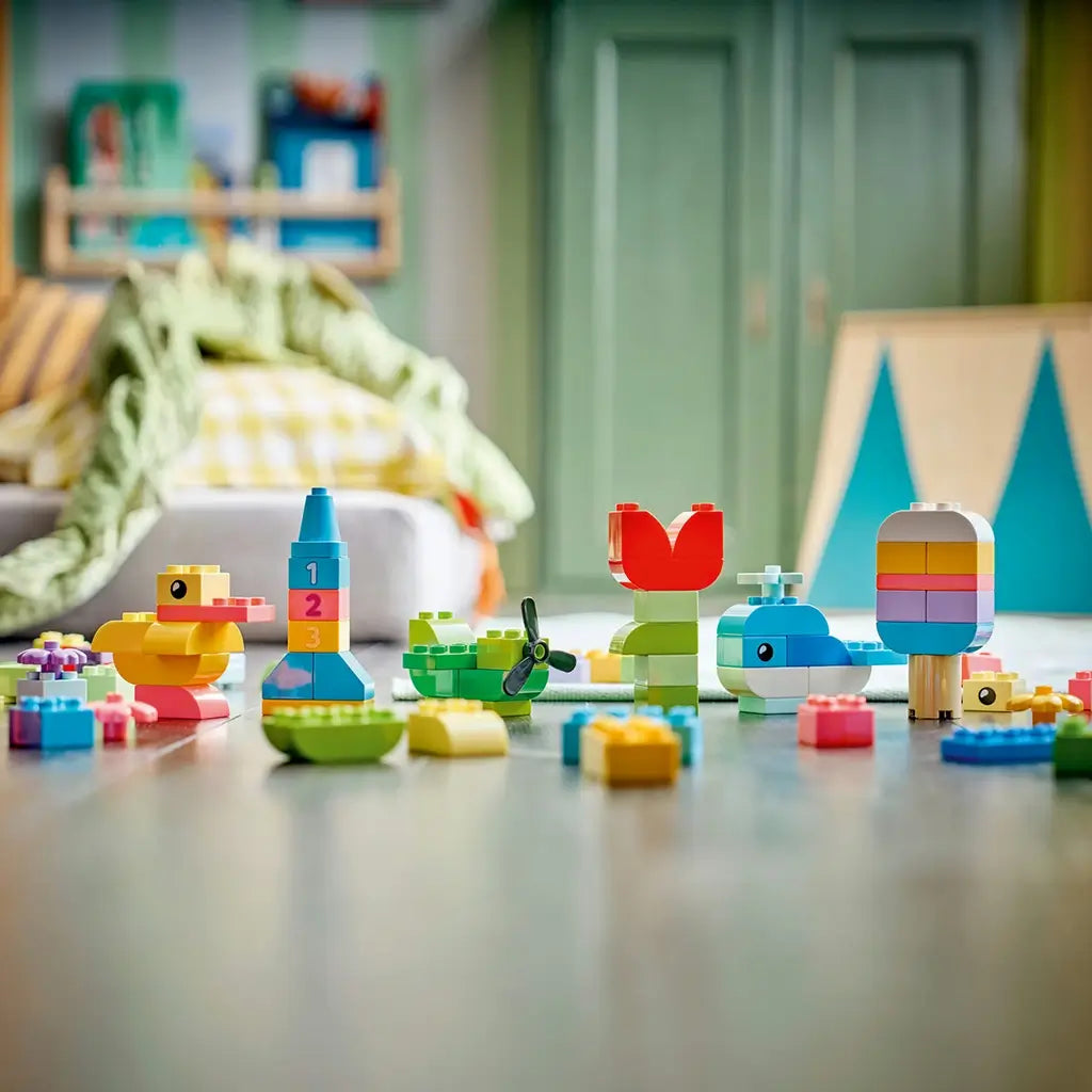 LEGO DUPLO Colorful Creative Box models use bright plastic bricks to form a duck, number tower, boat, flower and striped ice pop, displayed across a playroom floor.
