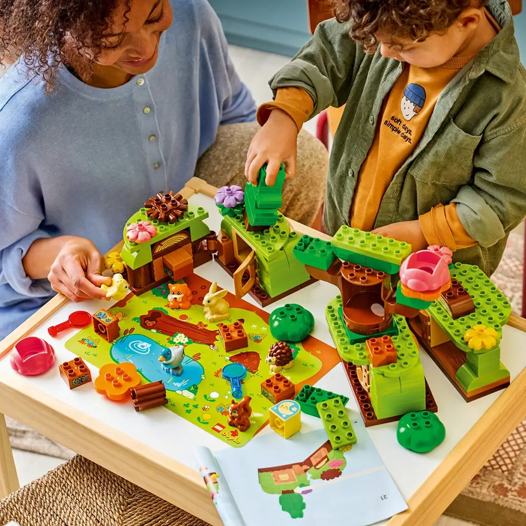 LEGO DUPLO Discovery Forest playset features green and brown plastic trees and habitats as a child builds beside an adult, with animal figures, flowers and a colorful play mat.