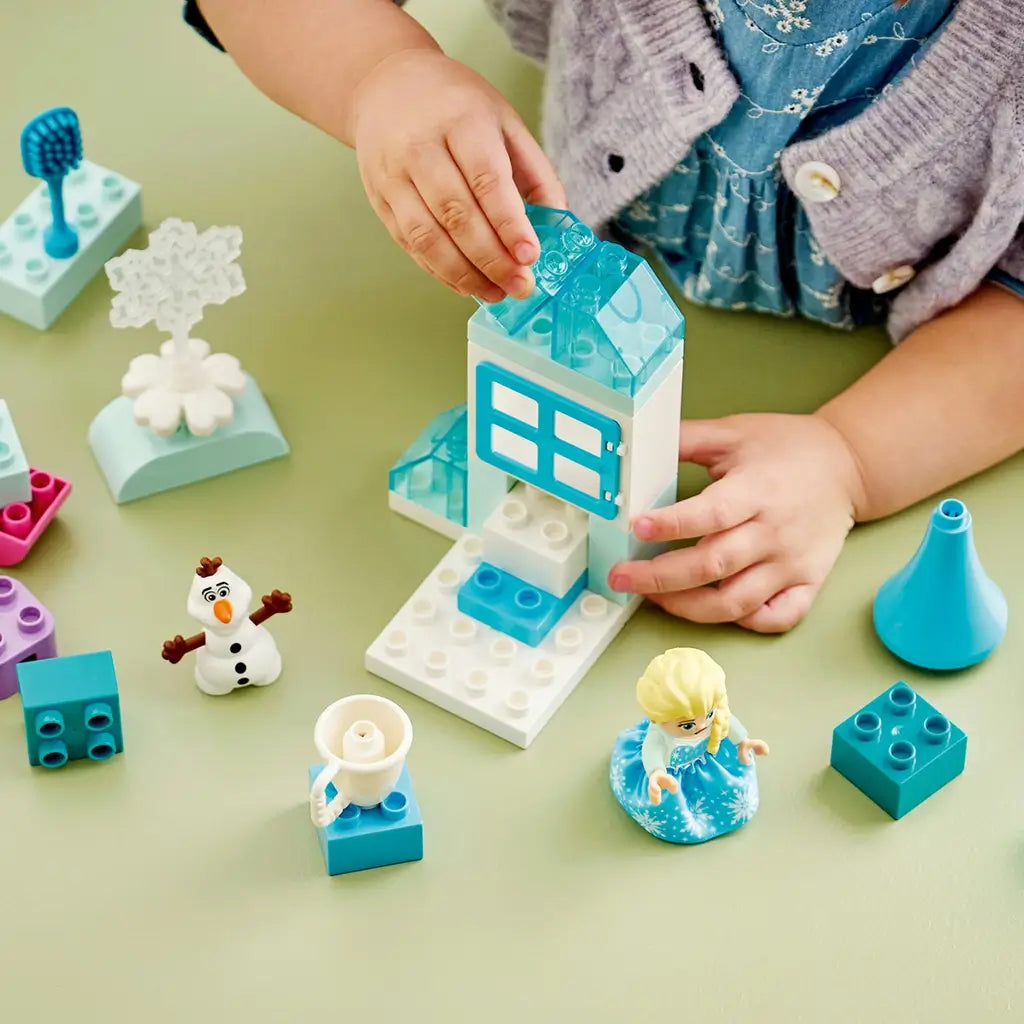 LEGO DUPLO Disney Frozen playset uses aqua, white and pastel plastic bricks as a child builds an icy house beside Elsa, Olaf, a cup and winter accessories.