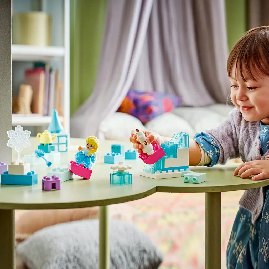 LEGO DUPLO Disney Frozen playset shows a child using aqua, white and pastel plastic bricks to play with Elsa and Olaf figures across a green table in a cozy room.