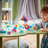 LEGO DUPLO Disney Frozen playset shows a child using aqua, white and pastel plastic bricks to play with Elsa and Olaf figures across a green table in a cozy room.