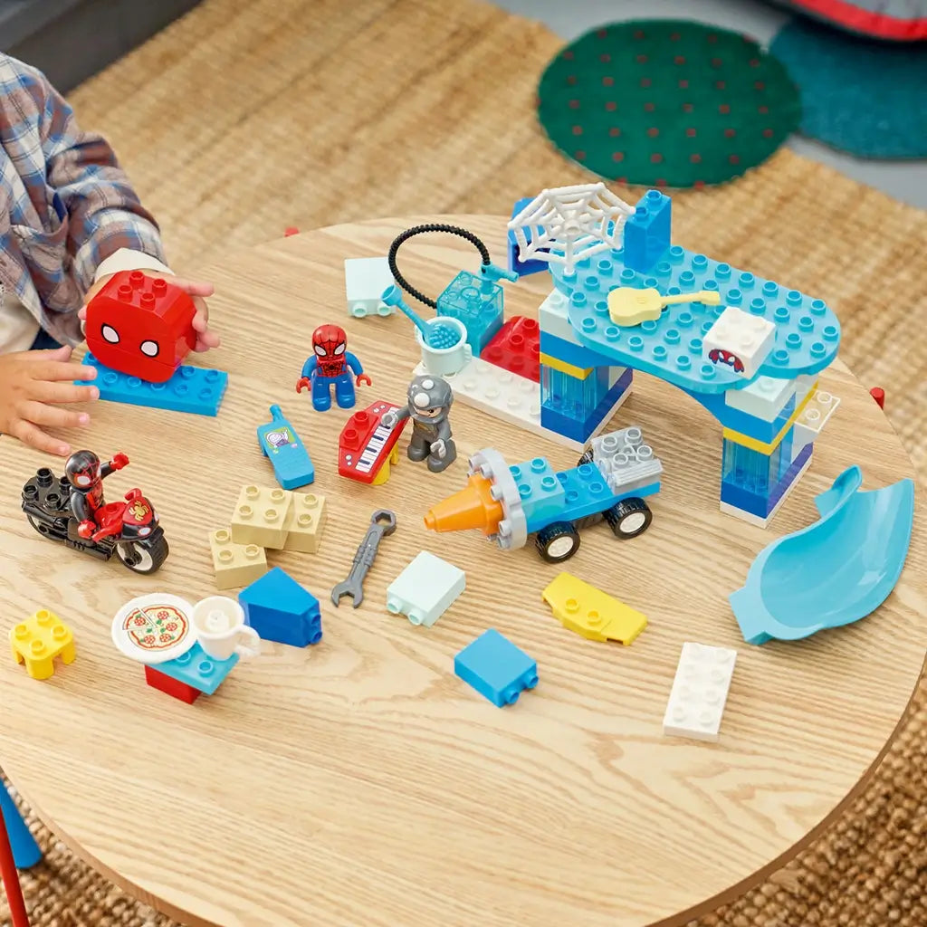 LEGO DUPLO Marvel Spidey Webquarters play scene shows blue and white plastic headquarters pieces, Spidey figures, vehicles and accessories spread across a wooden table.