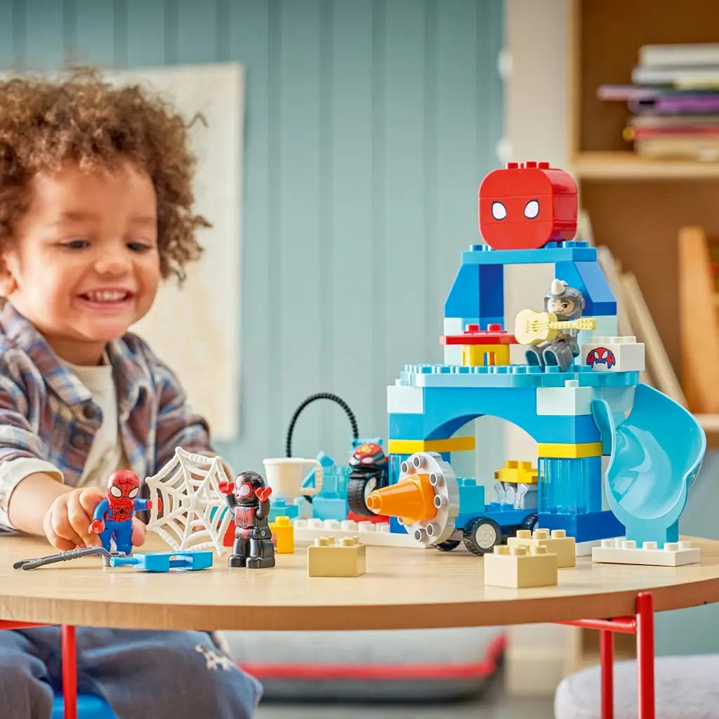 LEGO DUPLO Marvel Spidey Webquarters playset shows a child arranging Spidey figures beside a blue plastic headquarters with slide, drill vehicle and web on a table.