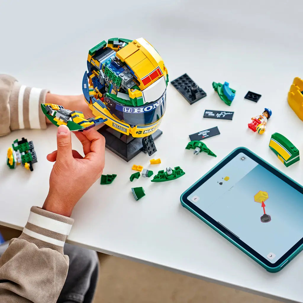 LEGO Ayrton Senna Helmet set 43024 is assembled from yellow, green and blue plastic bricks as a builder adds a side panel beside loose pieces and digital instructions.