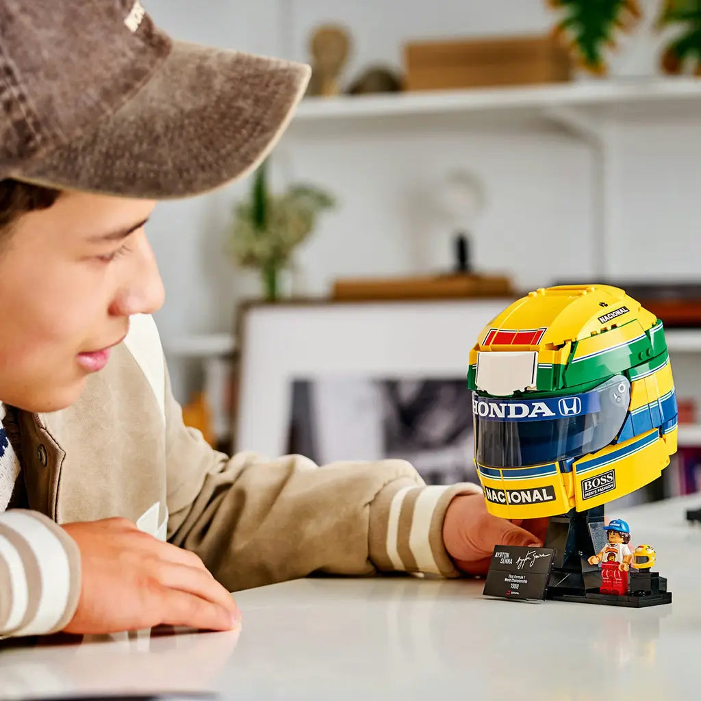 LEGO Ayrton Senna Helmet set 43024 displays a yellow, green and blue brick-built racing helmet on a black stand with plaque and minifigure as a young builder looks on.