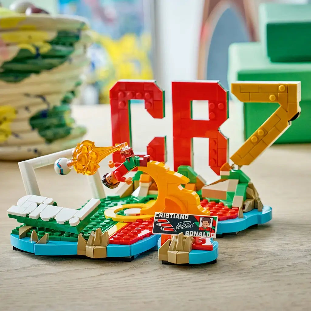 LEGO Cristiano Ronaldo soccer display built with red, green, gold and white plastic bricks, forming CR7 lettering, a goal frame and bicycle kick action scene on a neutral studio background.