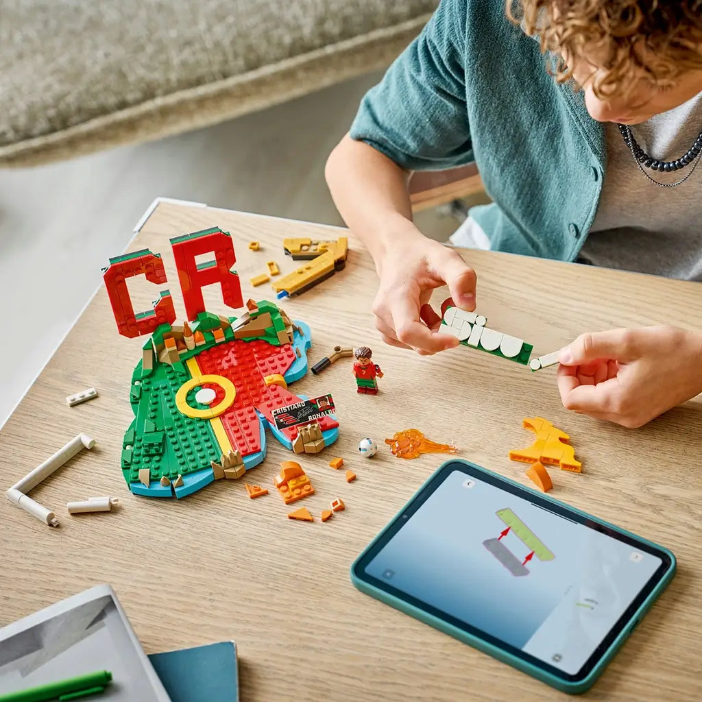 LEGO Cristiano Ronaldo soccer set in red, green, gold and white plastic bricks, partially assembled with loose components, minifigure and digital instructions on a wooden tabletop workspace.