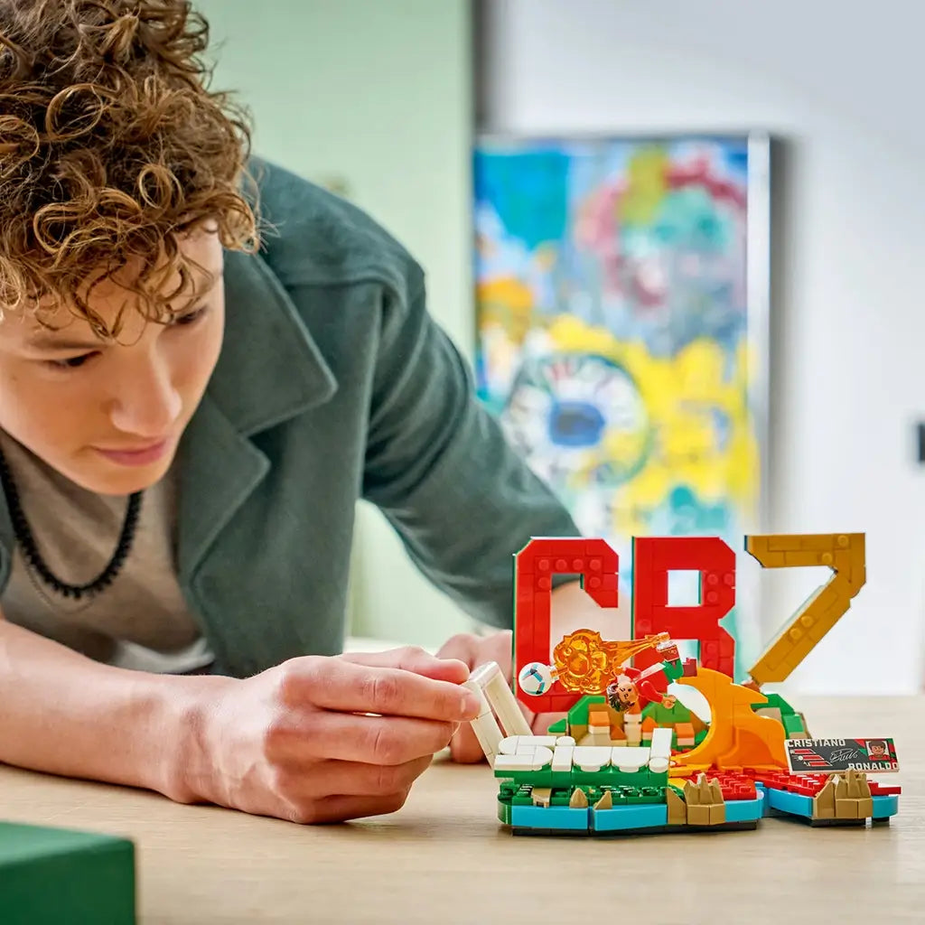 LEGO Cristiano Ronaldo soccer display in red, green, gold and white plastic bricks, compact CR7 highlight model being adjusted by a builder on a tabletop in a bright indoor environment.