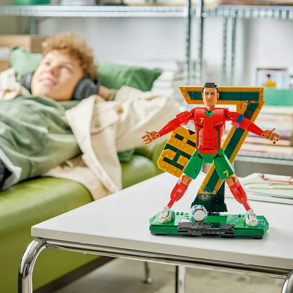 LEGO Cristiano Ronaldo soccer collectible features red, green, gold and black plastic brick construction displayed on a table with CR7 backdrop, while a fan relaxes nearby in a home setting.