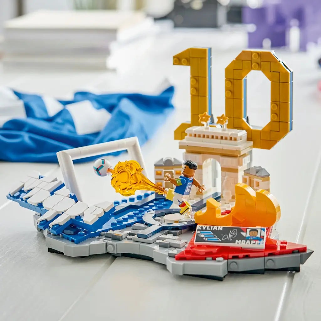 LEGO Kylian Mbappé soccer collectible features blue, white, gold and red plastic brick construction shaped as a football celebration scene with number 10, goal frame and landmark details, displayed on a tabletop.