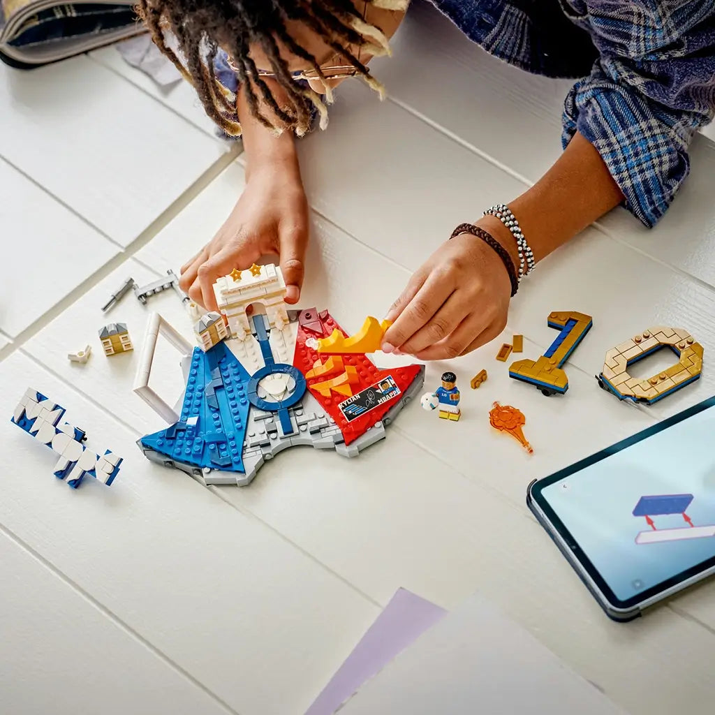LEGO Kylian Mbappé soccer building set features blue, white, gold and red plastic brick components forming a football highlights display, with a builder assembling pieces beside digital instructions.
