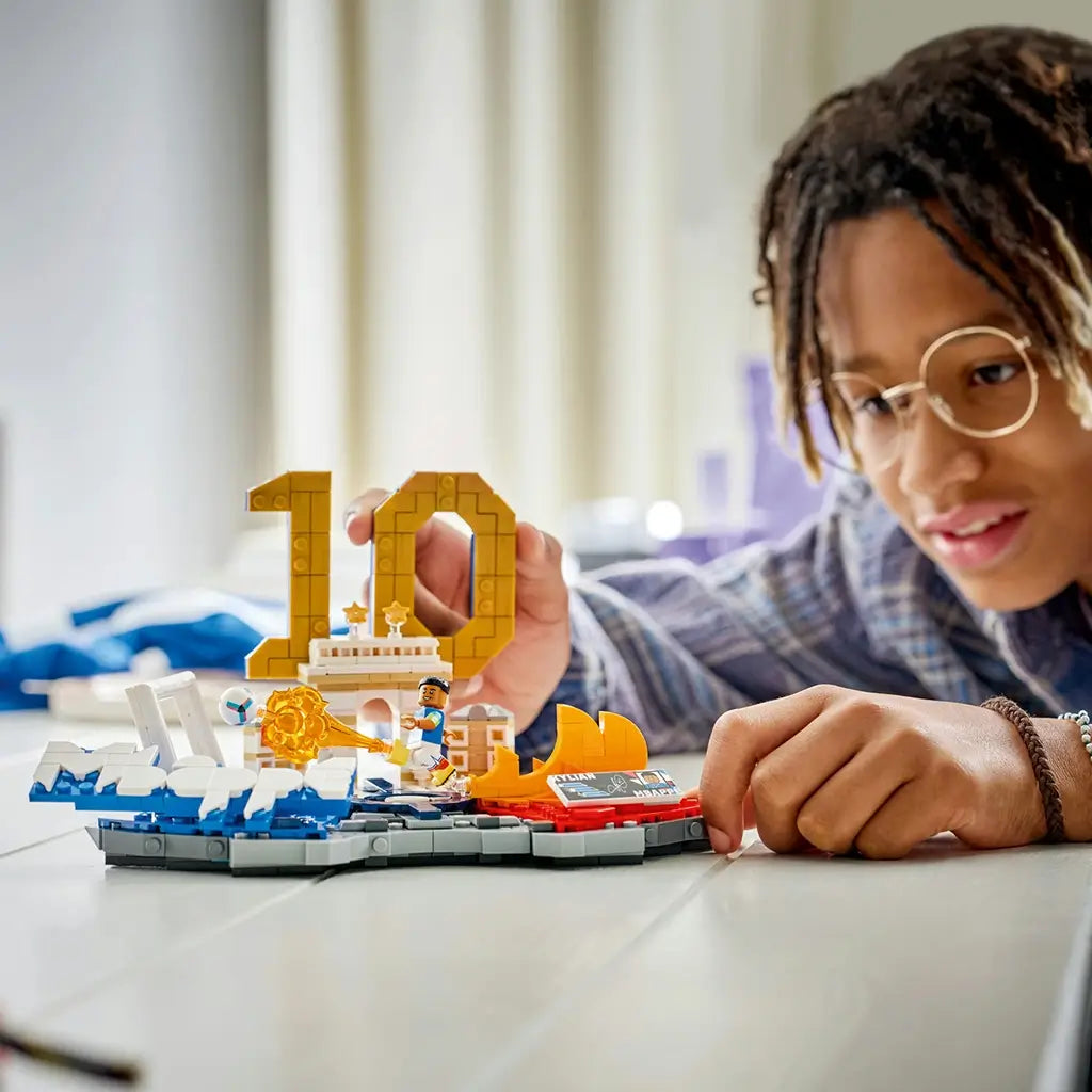 LEGO Kylian Mbappé soccer model features blue, white, gold and red plastic brick construction shaped as a football highlights display, with a builder interacting beside the number 10 backdrop and action scene.
