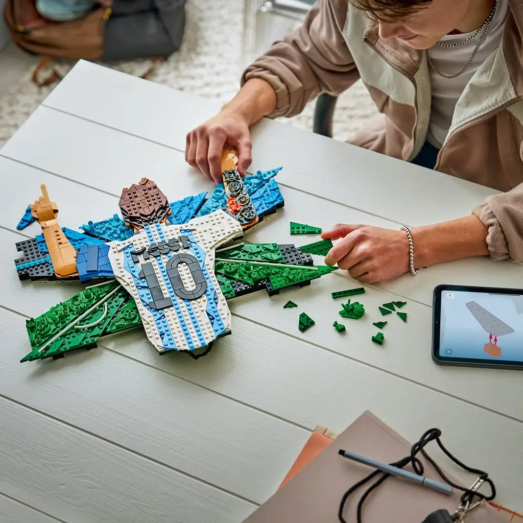 LEGO Lionel Messi building set features white, sky-blue, green and tan plastic brick sections forming a celebration wall display, shown during assembly on a tabletop beside digital building instructions.