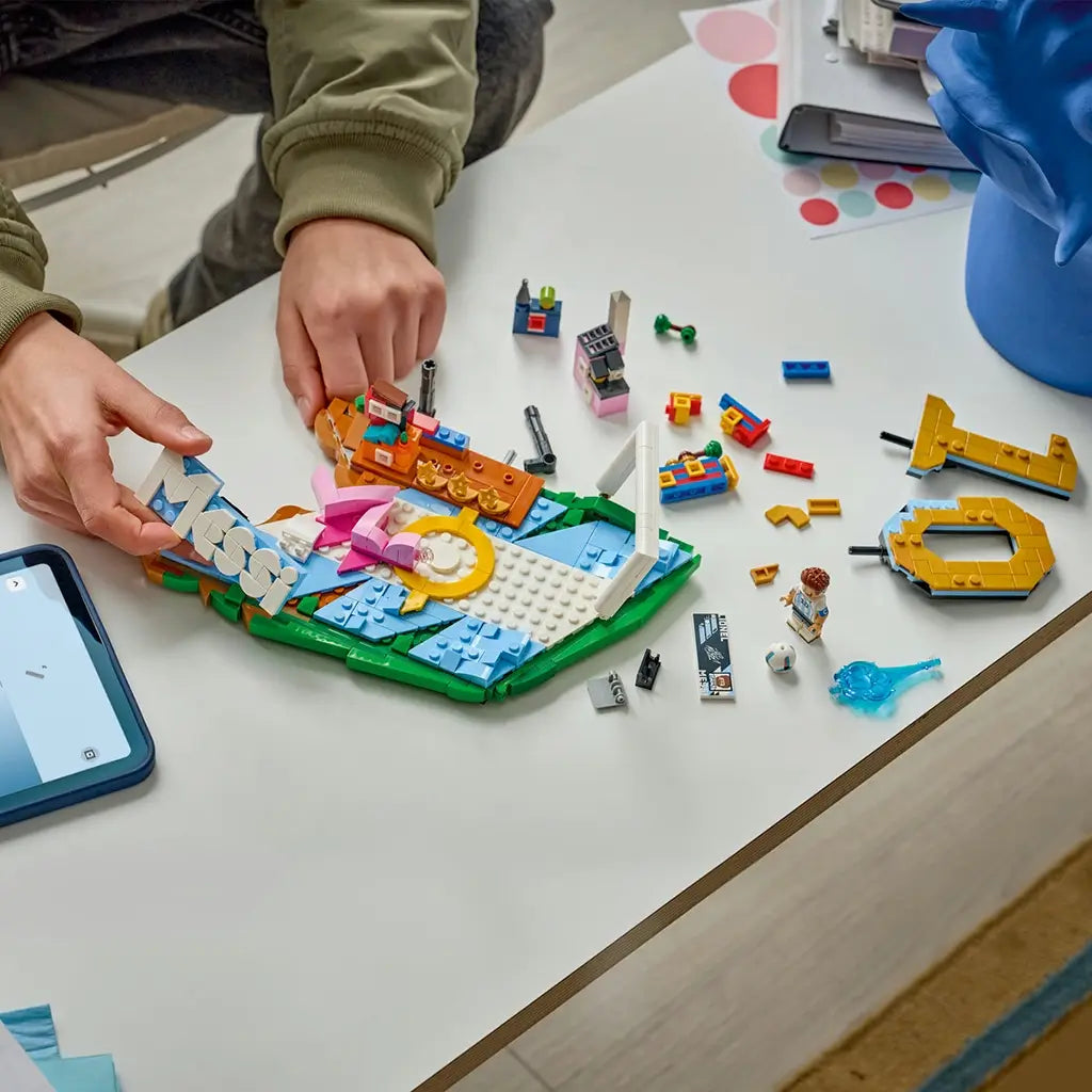 LEGO Lionel Messi soccer set in blue, green, white and gold plastic bricks, partially assembled with loose components, minifigure and digital instructions spread across a tabletop workspace.
