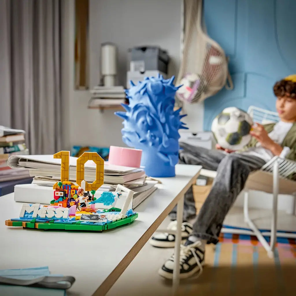 LEGO Lionel Messi soccer display in blue, green, white and gold plastic bricks, arranged as a number 10 highlight scene with goal frame and football effect, showcased on a desk in a home setting.