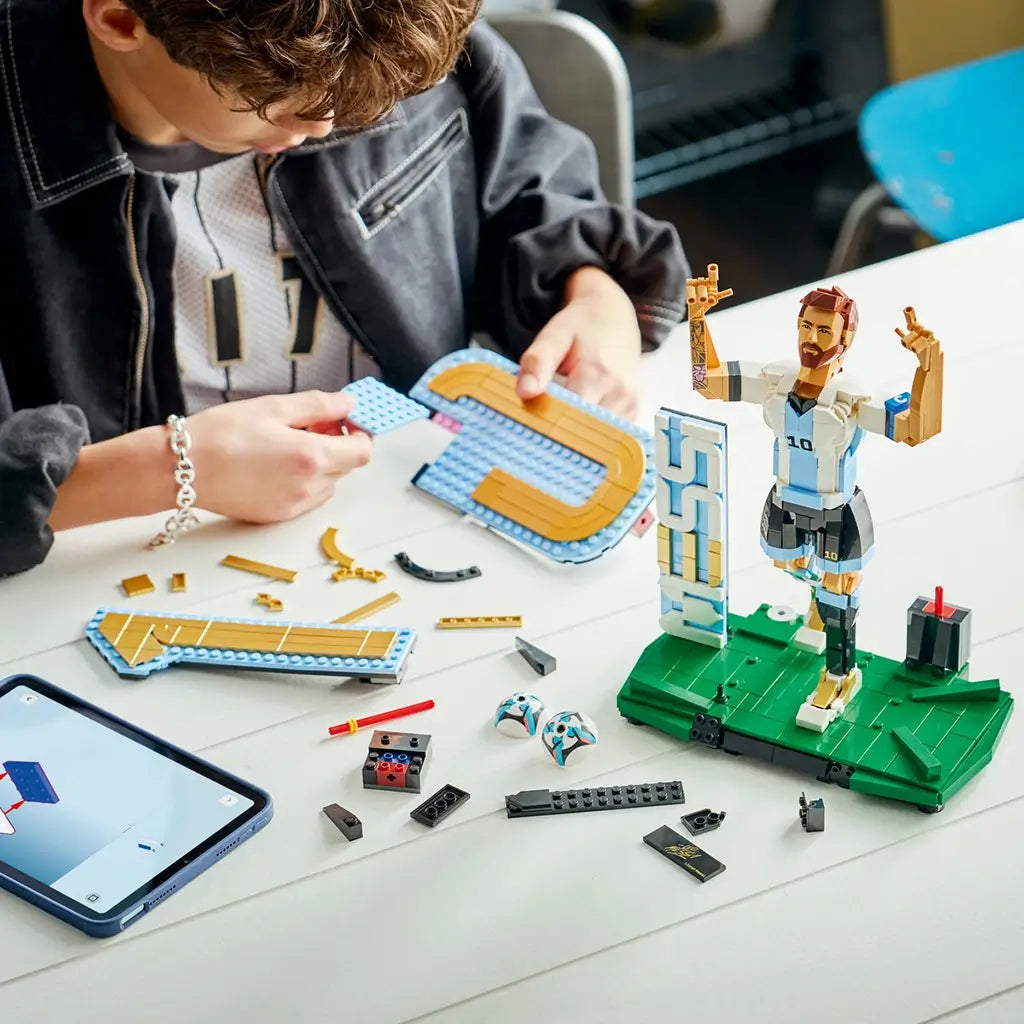 LEGO Lionel Messi soccer building set features light blue, white, black, gold and green plastic brick components being assembled beside digital instructions, with figure, backdrop and ball elements.