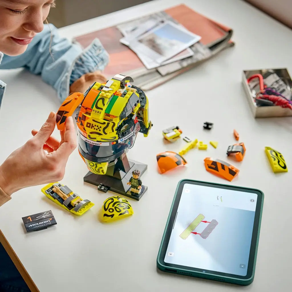 LEGO McLaren Formula 1 helmet building scene features yellow, orange and black plastic components, curved visor sections and assembly steps in progress on a tabletop beside digital instructions.