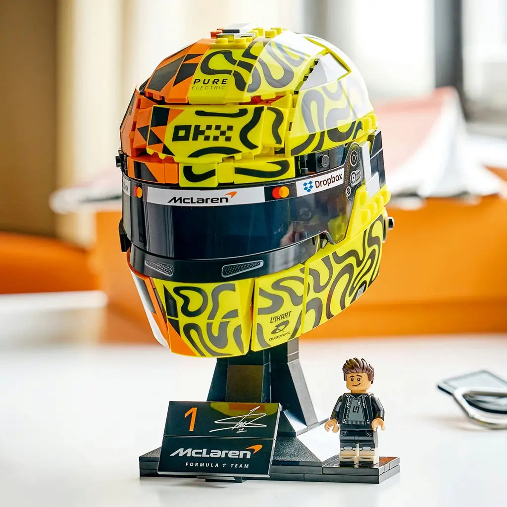LEGO McLaren Formula 1 helmet display features yellow, orange and black plastic panels with bold racing graphics, a dark visor and Lando Norris minifigure showcased on a stand in a bright room setting.