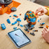LEGO McLaren Formula 1 helmet model features blue, orange and black plastic brick components being assembled on a tabletop, with removable panels, digital instructions and branded display elements nearby.