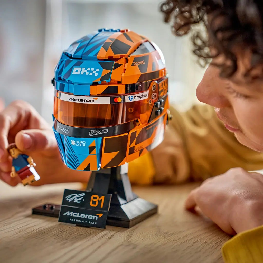 LEGO McLaren Formula 1 helmet collectible features blue, orange and black plastic brick construction styled after Oscar Piastri’s racing helmet, displayed on a stand while a builder holds the minifigure.