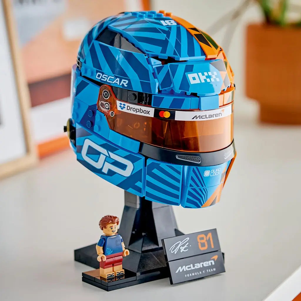 LEGO McLaren Formula 1 helmet display features blue, orange and black plastic brick construction shaped as Oscar Piastri’s racing helmet with reflective visor, mounted on a stand beside a minifigure.