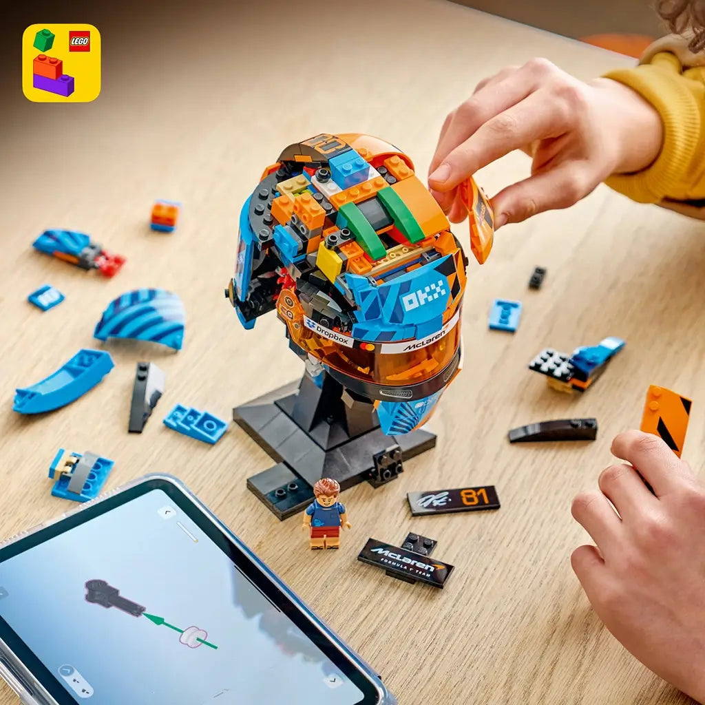 LEGO McLaren Formula 1 helmet building set features blue, orange and black plastic brick sections forming Oscar Piastri’s racing helmet, shown partially assembled beside digital instructions and loose pieces.