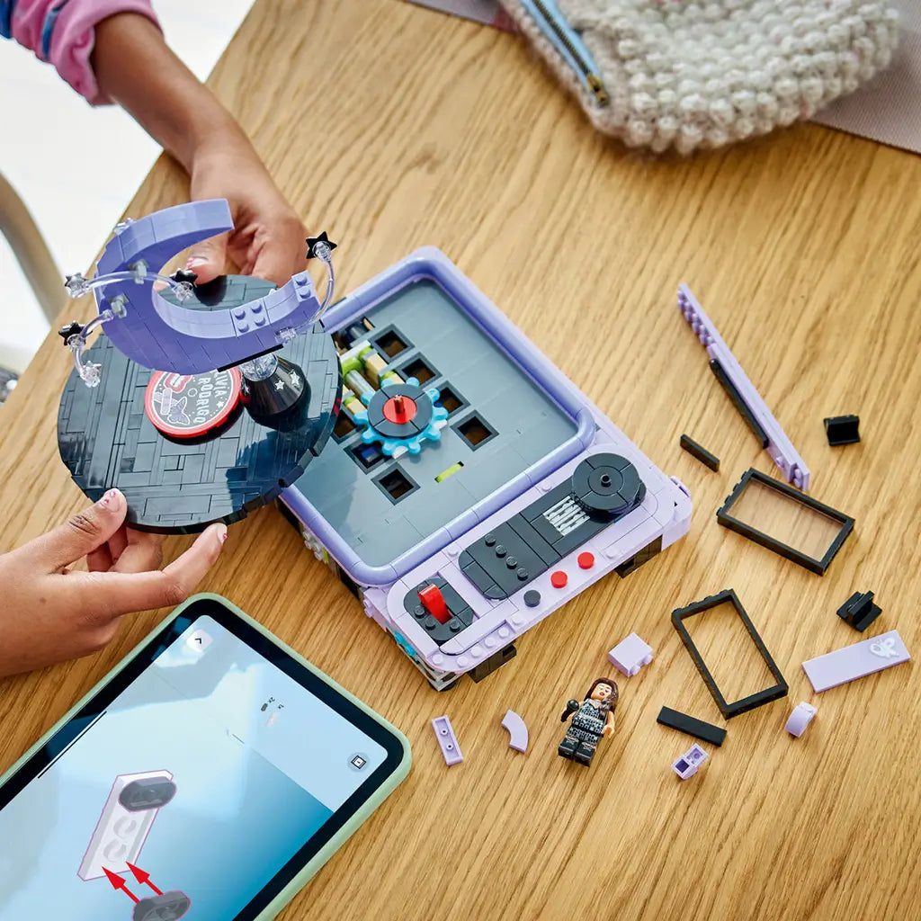 LEGO Olivia Rodrigo Concert Moon in lavender, black and gray bricks is being assembled on a wood table, with the crescent moon stage lifted beside digital instructions.