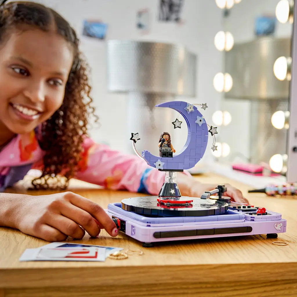 LEGO Olivia Rodrigo Concert Moon in lavender and black bricks forms a turntable and crescent moon stage as a smiling builder adjusts a control on a wood vanity table.
