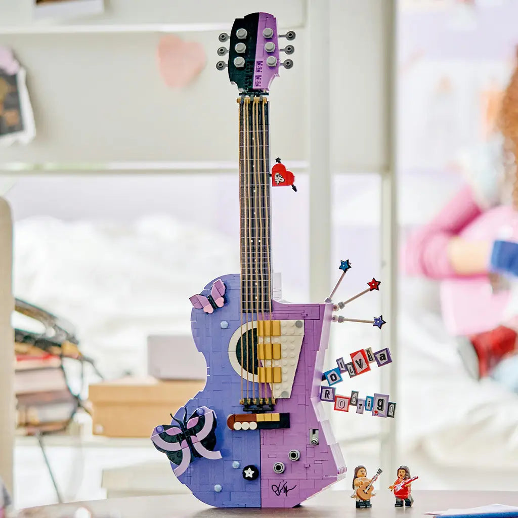 LEGO Olivia Rodrigo Dual Guitar in lavender, black and white bricks stands as a tall music display with butterfly, star accents and two minifigures in a bright room.