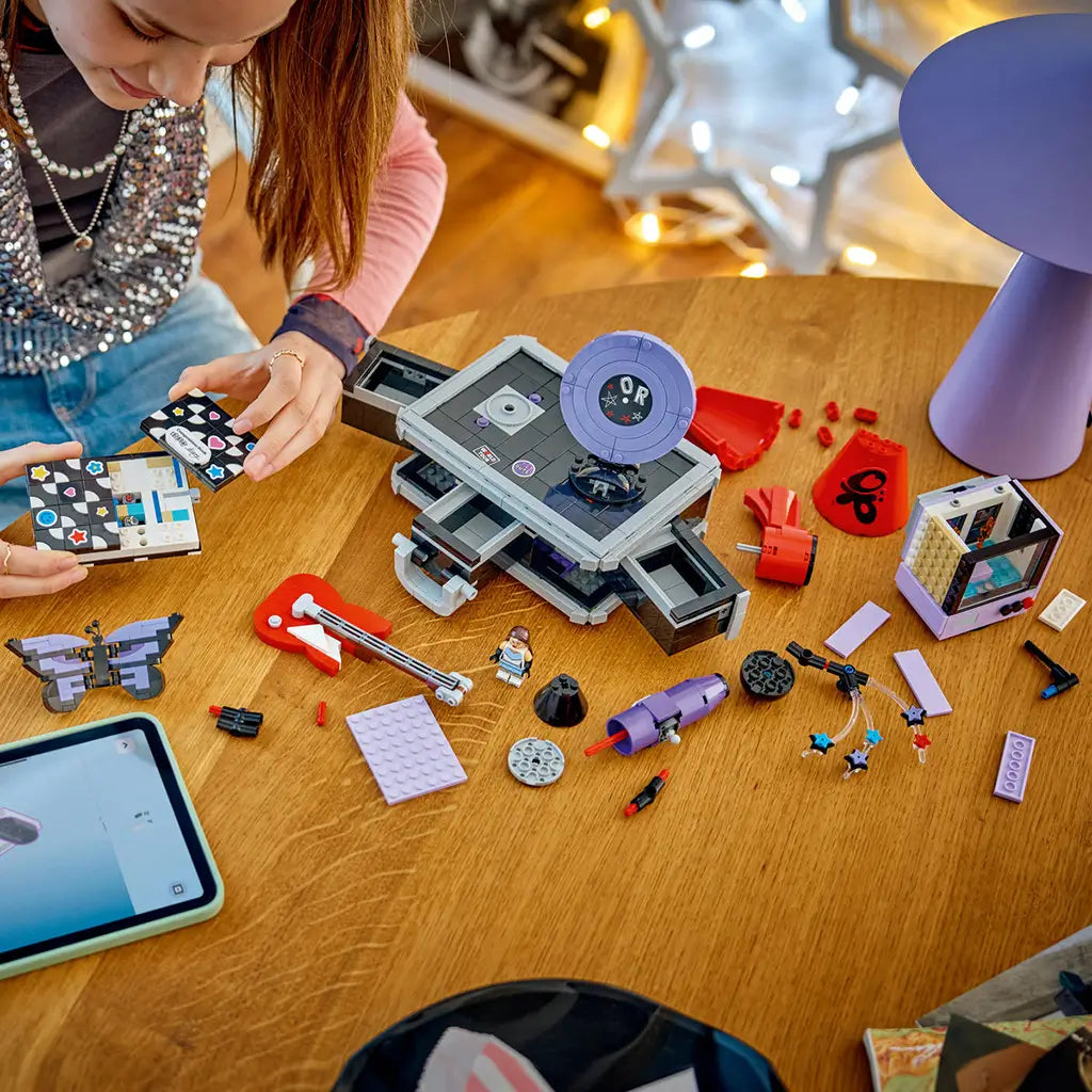 LEGO Olivia Rodrigo Secret Storage pieces in black, gray, lavender and red cover a wood table as a builder assembles the music case beside digital instructions.